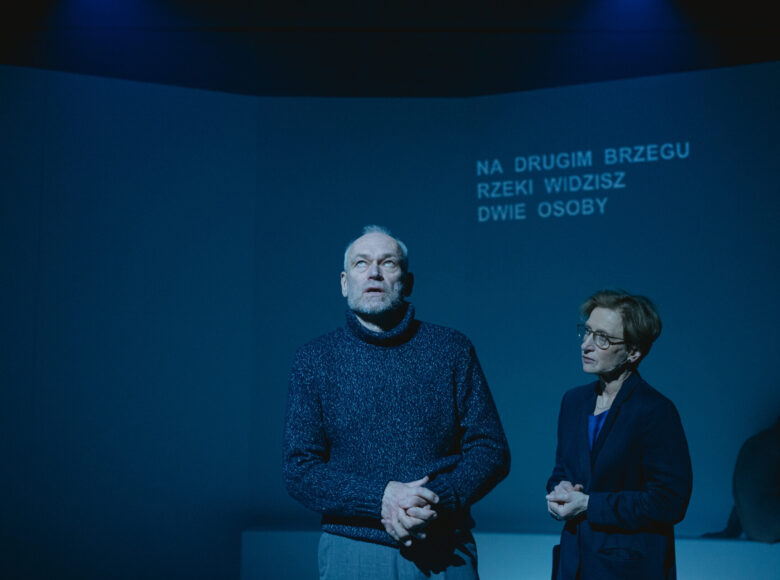 Kobieta i mężczyzna stoją na tle ściany. Mężczyzna ma oczy zwrócone do góry, kobieta patrzy na niego. W tle za nimi napis „ na drugim brzegu rzeki widzisz dwie osoby”.