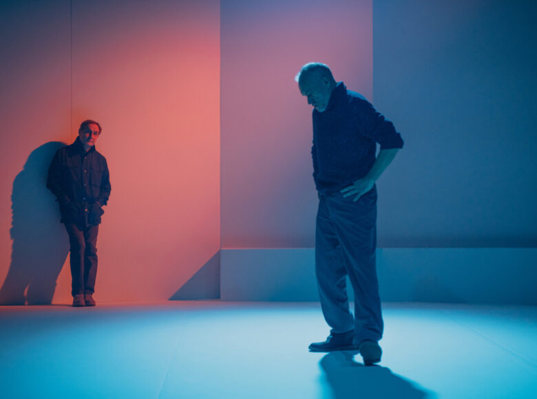 Dwóch mężczyzn stoi na tle ściany oświetlonej na brudnoróżowy kolor. Pierwszy bliżej kadru ma ręce oparte na biodrach, patrzy na podłogę. Drugi, oparty na ścianę, patrzy na pierwszego.