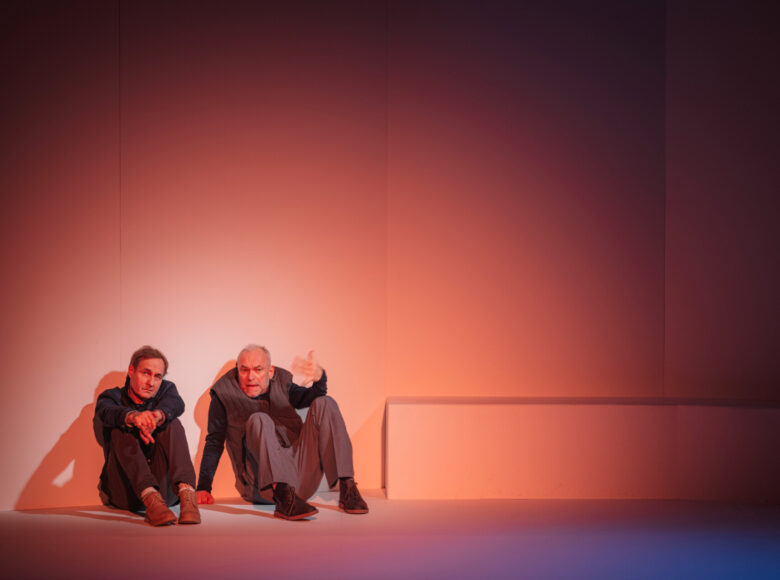Dwóch mężczyzn siedzi na podłodze, opartych o ścianę oświetloną na różowo-pomarańczowo. Jeden wydaje się coś opowiadać, jedną rękę ma wyciągniętą przed siebie.