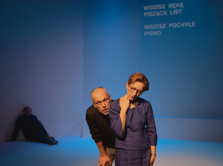 Na zdjęciu trzy osoby. Na pierwszym planie stoi kobieta, jedną rękę trzyma na szyi. Za jej plecami stoi mężczyzna, wydaje się coś mówić. W tle, częściowo widoczny mężczyzna siedzi na podłodze, oparty o ścianę. Ściana oświetlona na niebiesko, na niej napis „widzisz rękę piszącą list. Widzisz pochyłe pismo”.