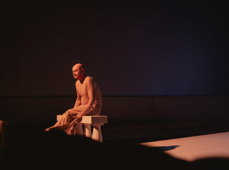 W centrum zdjęcia mężczyzna siedzi na białym, niskim postumencie z 3 kolumnami. Jest lekko oświetlony, dookoła niego scena ciemna.