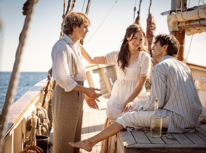 Kolorowa fotografia przedstawia trzy osoby (od lewej: mężczyzna, kobieta, mężczyzna) na statku. Ubrani są w jasne, lekkie ubrania i rozmawiają. Mężczyzna po lewej stoi, natomiast reszta siedzi na deskach. Przed jednym z mężczyzn stoi szklana butelka.