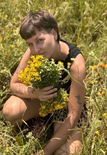 Zdjęcie przedstawia osobę na łące. Siedzi ona wśród zielonych traw i trzyma bukiet z polnych, żółtych kwiatów. Osoba patrzy się prosto w obiektyw, ma fryzurę typu mullet, zielone oczy, kilka kolczyków i tatuaży.