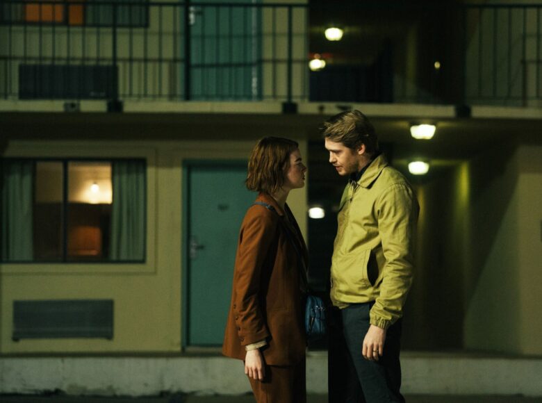 Kolorowy fotos z filmu przedstawia kobietę i mężczyznę stojących przed budynkiem wieczorem. Oboje stoją bardzo blisko siebie i skierowani są ku sobie. Kobieta ma na sobie brązową marynarkę oraz brązowe spodnie, a mężczyzna jasnobrązową kurtkę oraz czarne spodnie.