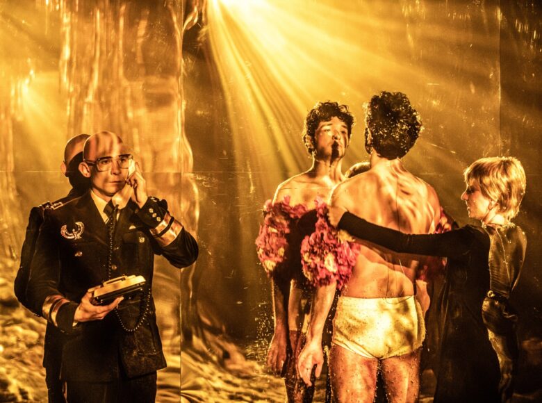 Grupa osób stoi przed lustrzaną ścianą. Po lewej stronie mężczyzna w czarnym uniformie, w rękach trzyma stary telefon stacjonarny, a słuchawkę na kablu przy uchu. Drugi mężczyzna w samych slipach i czerwonym szalu z piór stoi twarzą do lustra, za ramiona trzyma go kobieta, stojąca za nim.