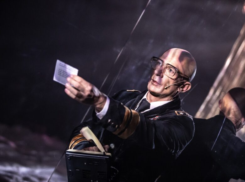 Zbliżenie na mężczyznę, widzianego od pasa w górę. Ubrany w czarny uniform, w wyciągniętej przed sobą dłoni trzyma niewielką kartkę.