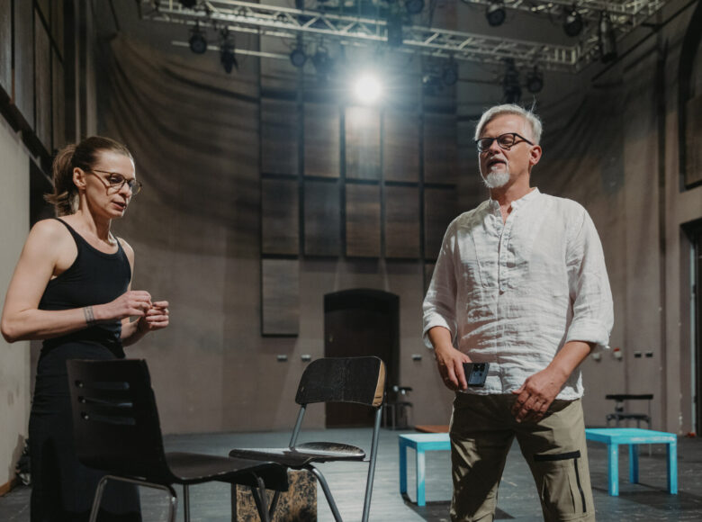 Kolorowe zdjęcie przedstawia od lewej aktora i aktorkę. Pomiędzy nimi stoją dwa czarne krzesła. Kobieta ubrana jest na czarno, wzrok skierowany ma do dołu, a mężczyzna ubrany jest w lnianą, jasną koszulę i jasnobrązowe spodnie. W dłoni trzyma telefon, a oczy ma zamknięte.