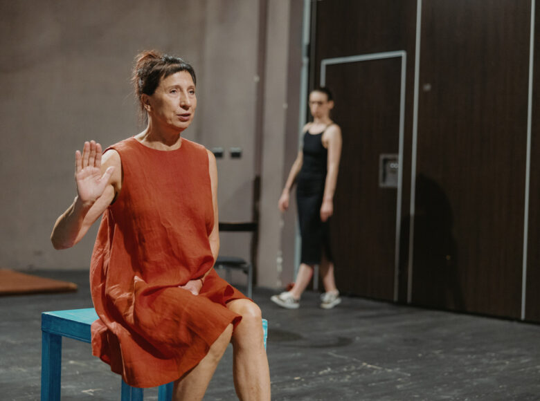 Na pierwszym planie fotografii znajduje się kobieta w pomarańczowej sukience, która siedzi na niebieskim stoliczku i trzyma jedną rękę w górze. W tle, za nią przechadza się kobieta w czarnej sukience, która zerka na tą z pierwszego planu.