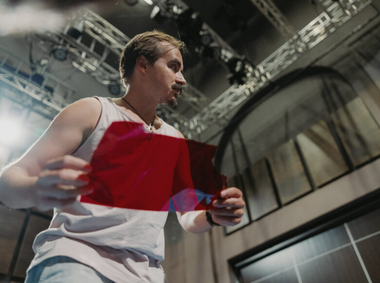 Zdjęcie ukazuje aktora z perspektywy od dołu. Ubrany jest w jasną koszulkę, a w dłoni trzyma czerwony materiał.