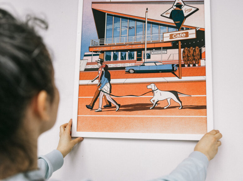 Fotografia przedstawia osobę trzymającą obraz przy ścianie. Na obrazie para osób wyprowadza psa na spacer. Na obrazku przeważają kolory: pomarańczowy i niebieski. Para z psem przechadza się na tle budynku, w którego szybie odbija się startujący samolot.