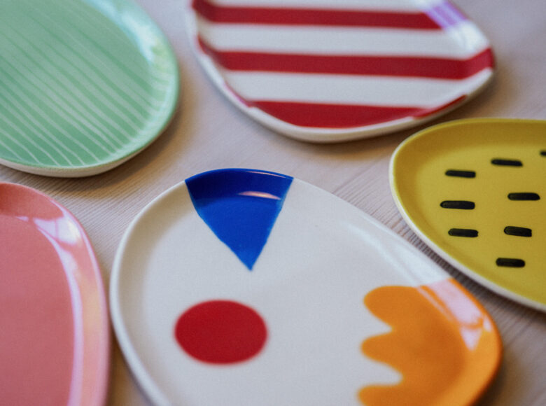 Kolorowe zdjęcie przedstawia talerze o różnych wzorach i barwach. Talerze od lewej: różowy, ciemnozielony w jasnozielone paski, z czerwonym kółkiem, niebieskim trójkątem oraz pomarańczową plamą, w białe i czerwone pasy, zółtry w czarne, krótkie paski.