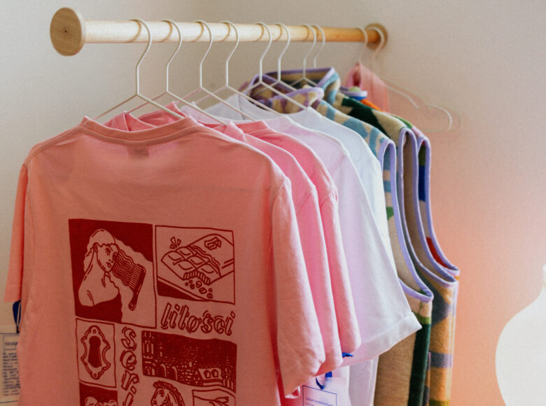 Kolorowa fotografia przedstawia koszulki powieszone na drewnianym wieszaku jedna za drugą. Pierwsza jest koloru brzoskwiniowego i ma na sobie grafiki kobiety czeszącej się grzebieniem, czekolady, otworu na klucz, budynków, konia, oraz napisy "litości" i "serio".