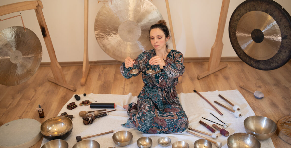 Zdjęcie przedstawia kobietę grającą na instrumentach. Kobieta ubrana jest w niebiesko brązową sukienkę. Dziewczyna siedzi wśród swoich instrumentów, a na twarzy widać jej skupienie nad muzyką.