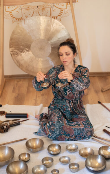 Zdjęcie przedstawia kobietę grającą na instrumentach. Kobieta ubrana jest w niebiesko brązową sukienkę. Dziewczyna siedzi wśród swoich instrumentów, a na twarzy widać jej skupienie nad muzyką.