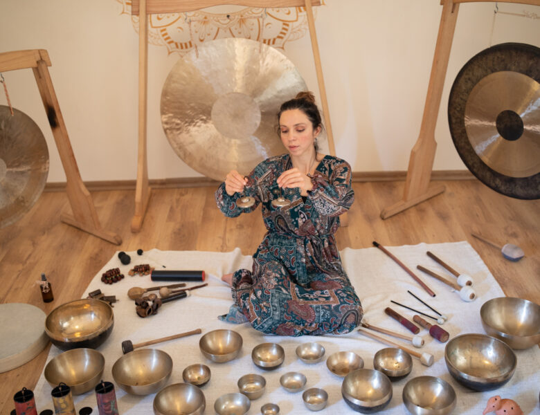 Zdjęcie przedstawia kobietę grającą na instrumentach. Kobieta ubrana jest w niebiesko brązową sukienkę. Dziewczyna siedzi wśród swoich instrumentów, a na twarzy widać jej skupienie nad muzyką.