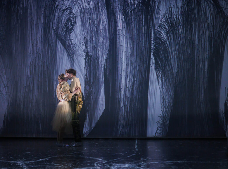 Scena w teatrze. Dziewczyna i chłopak obejmują się i całują w usta. Za nimi ciemna ściana ozdobiona abstrakcyjnymi rysunkami, przypominającymi kształty drzew.