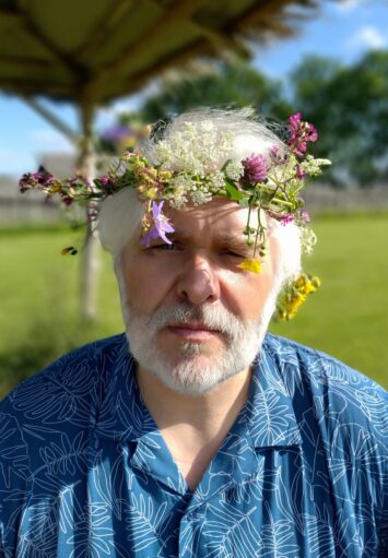 Kolorowe zdjęcie przedstawia mężczyznę o siwych włosach i brodzie patrzącego prosto w obiektyw. Ubrany jest w niebieską koszulę z wyszytymi na biało liśćmi. Na głowie ma kolorowy wianek z kwiatów.