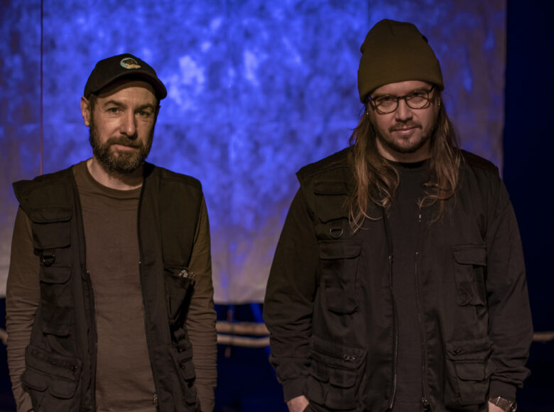 Dwóch mężczyzn stoi na tle ekranu oświetlonego na niebiesko, patrząc w obiektyw.