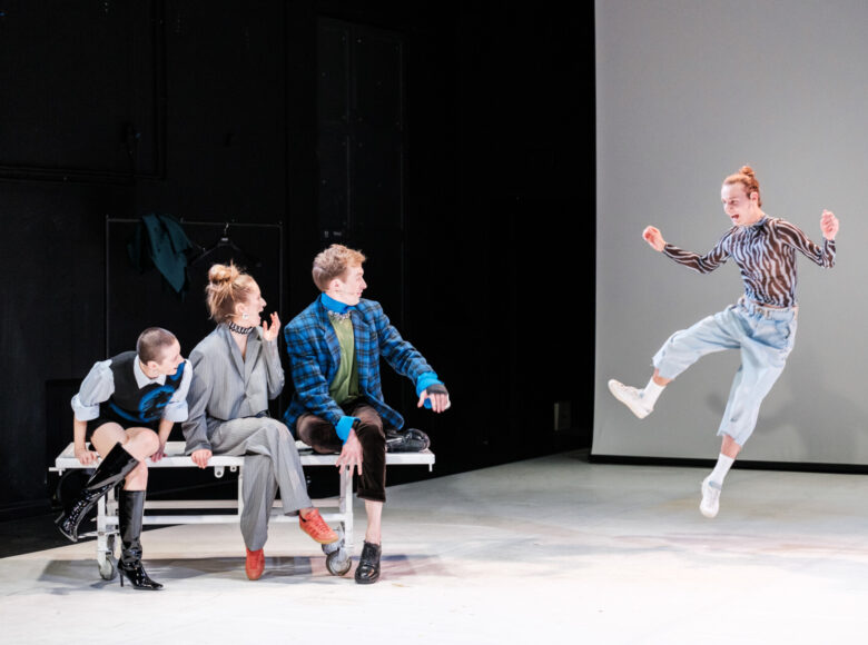 Scena grupowa. Po lewej stronie kadru na ławce siedzą dwie kobiety i mężczyzna. Patrzą się w prawą stronę kadru, gdzie widoczny jest jeden mężczyzna w wyskoku.