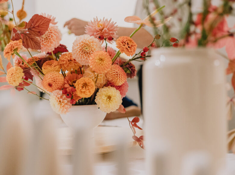 Kolorowe zdjęcie przedstawia białe donice, w których znajdują się beżowe, pomarańczowe i bladoróżowe kwiaty. Gdzieniegdzie przebijają się zielone łodygi.