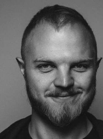 Czarno - białe zdjęcia przedstawia portret uśmiechniętego mężczyzny o krótkich włosach i brodzie.