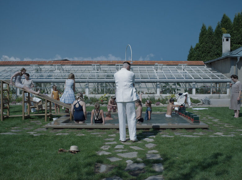 Kadr z filmu. Na zewnątrz. Podwórko z niewielkim basenem. W tle szklarnia i mały budynek/ W basenie i na zjeżdżalni obok grupa dzieci. Wkoło basenu kilkoro dorosłych – kobiety w sukienkach i obrócony tyłem do nas mężczyzna w białym garniturze.