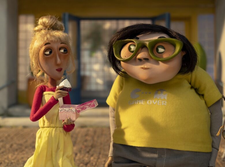 Kolorowa grafika przedstawia dwie postaci. Po lewej szczupła, o blond włosach zawiązanych w kok kobieta, ubrana jest w żółtą sukienkę, a pod nią ma czerwoną bluzkę z długim rękawem, je ona babeczkę spoglądając na postać po prawej stronie. Jest ona nieco większa i ma na sobie żółtą koszulkę z napisem "game over" oraz zielone okulary. Postać ma krótkie, czarne włosy.