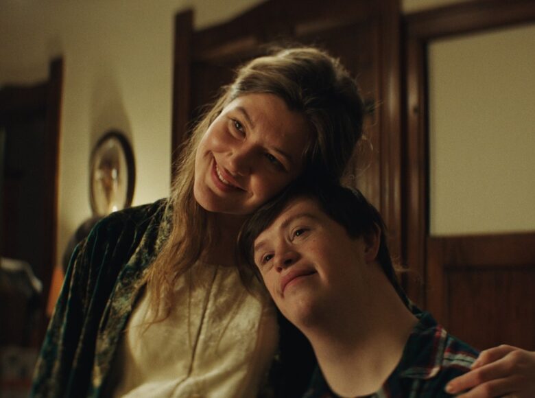 Zdjęcie przedstawia portret dwóch osób. Wyższa kobieta o długich blond włosach uśmiecha się i patrzy w dal obejmując chłopaka z zespołem downa za ramię. Mężczyzna również jest uśmiechnięty i patrzy w tą samą stronę co ona. Oboje opierają się o siebie głowami.