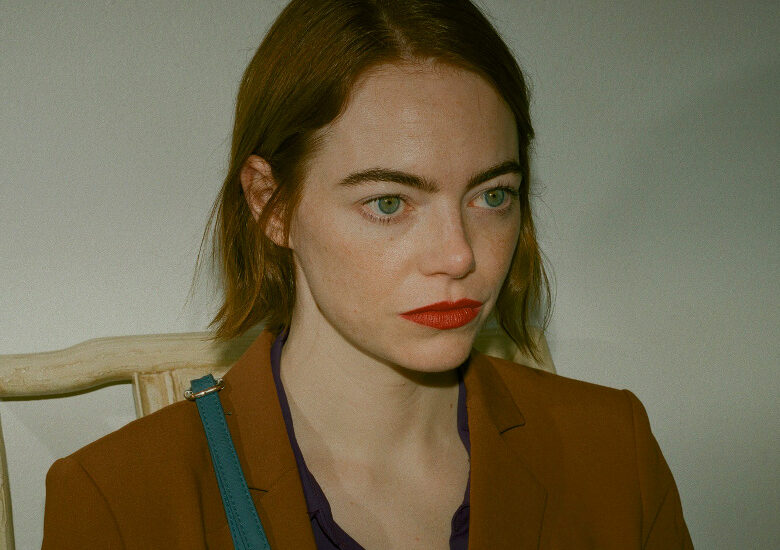Kadr z filmu. Młoda kobieta o zielonych oczach i umalowanych na czerwono ustach, w brązowej marynarce, siedzi na krześle na tle białej ściany.