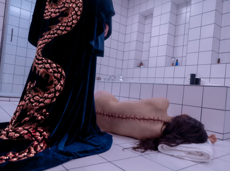 Kadr z filmu. Wnętrze łazienki wyłożonej białymi kafelkami. Na podłodze obok wanny leży naga kobieta. Widać jej plecy ze skórą rozciętą i zszytą wzdłuż całego kręgosłupa. Nad nią stoi postać w długim niebieskim szlafroku z wyhaftowanym złotym chińskim smokiem.