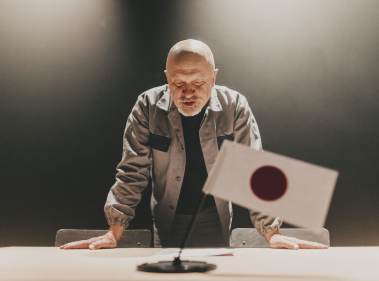 Scena teatralna. Za stołem stoi mężczyzna w szarej kurtce. Na stole stoi mała flaga Japonii.