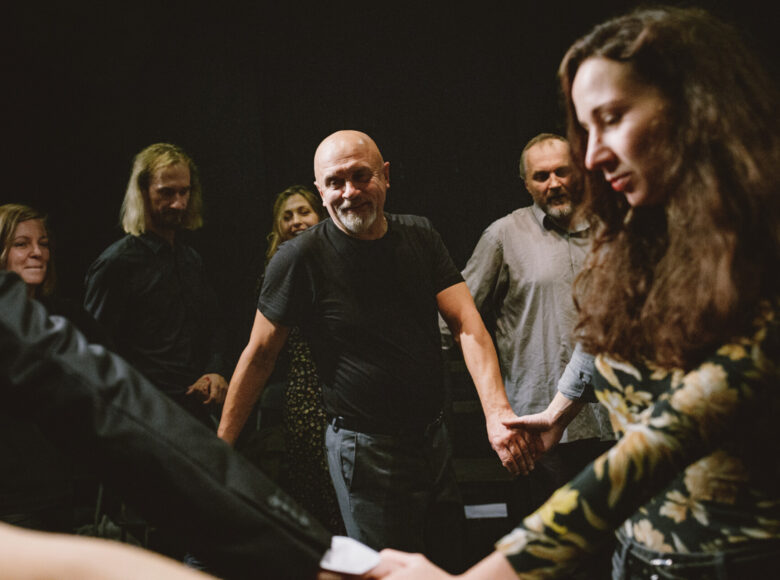 Przedstawienie teatralne. Na ciemnym tle grupa tańczących osób trzymających się za ręce.