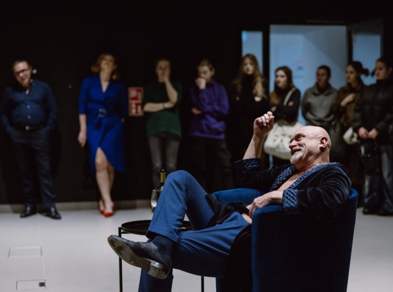 Przedstawienie teatralne. Na pierwszym planie aktor siedzi w fotelu w swobodnej pozie. Ubrany jest w niebieski kostium teatralny. W tle osoby z publiczności.