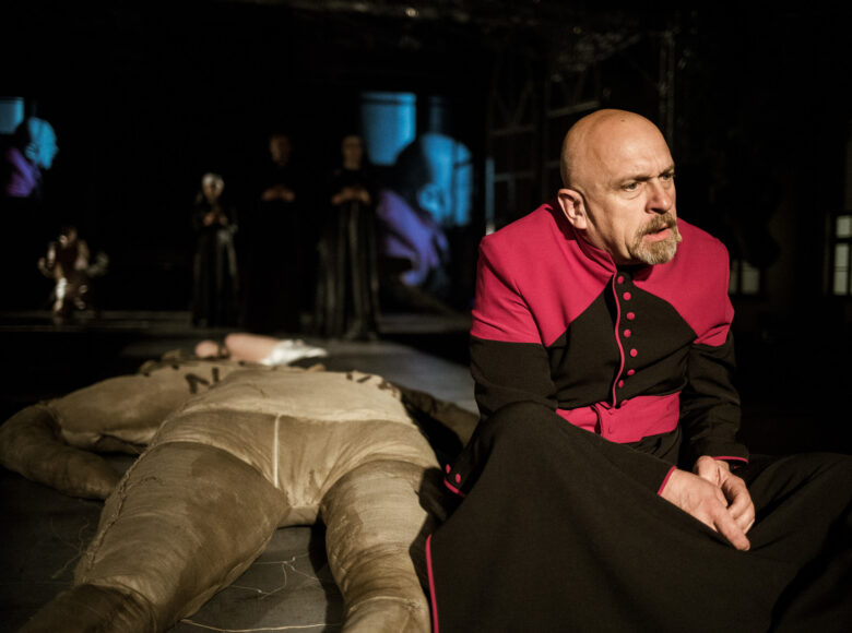 Scena teatralna. Na pierwszym planie mężczyzna w sutannie biskupiej.