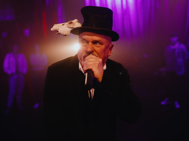 Przedstawienie teatralne. Na pierwszym planie mężczyzna w czarnym garniturze i czarnym kapeluszu. Do kapelusza przyczepiona mała czaszka zwierzęcia.