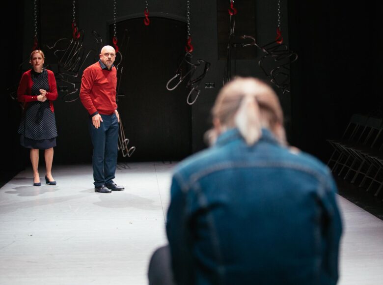 Na scenie teatralnej kobieta i dwóch mężczyzn. Kobieta i jeden z mężczyzn stoją, drugi mężczyzna siedzi.