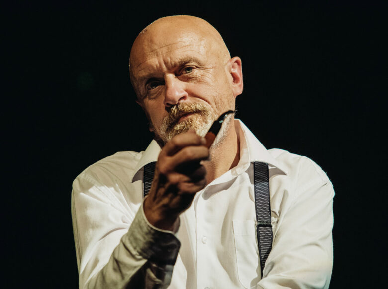 Przedstawienie teatralne. Na czarnym tle elegancko ubrany mężczyzna z fajką w dłoni.