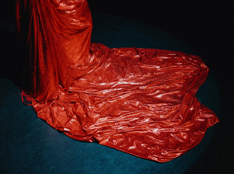 Scena teatralna. Czarne tło i czarna podłoga. Na środku czerwona udrapowana tkanina zwieszająca się z góry i rozkładająca się na podłodze.