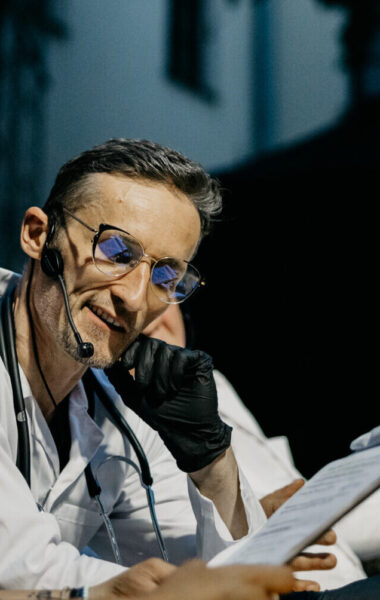 Przedstawienie teatralne. Zbliżenie na kobietę i mężczyznę w kitlach lekarskich, siedzących przy stole. Oboje patrzą na zapisaną kartkę, którą trzyma w ręku kobieta. W tle dwóch innych mężczyzn w kitlach lekarskich rozmawia ze sobą gestykulując. Jeden z nich siedzi, drugi stoi.