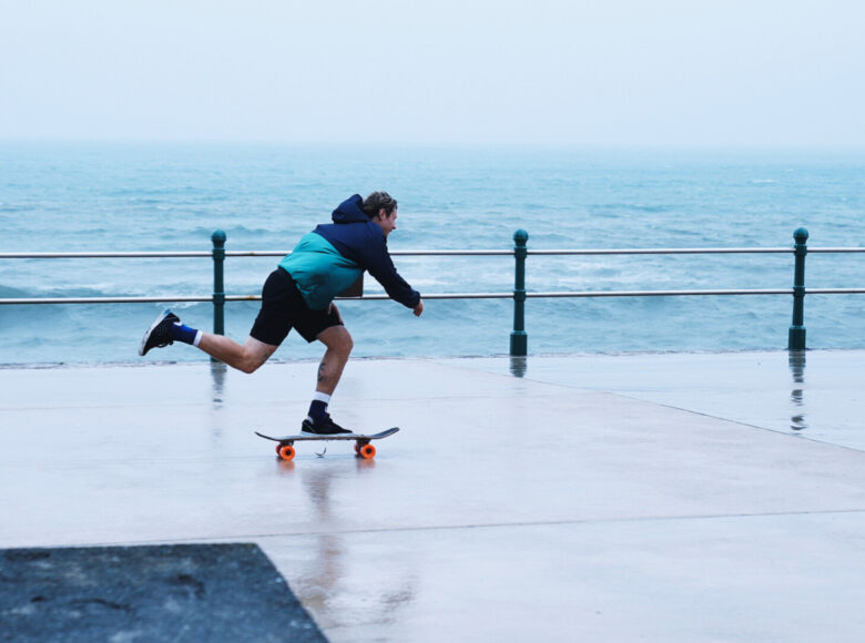 Pochmurny dzień. Na pierwszym planie most, za nim falujące morze. Na moście mężczyzna jadący na deskorolce.