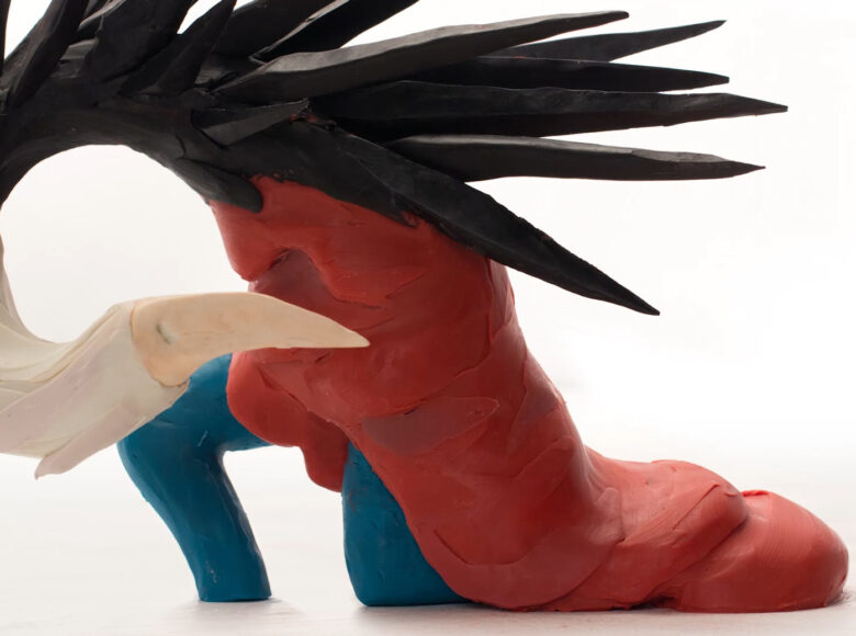 Na białym tle abstrakcyjny kształt ulepiony z plasteliny, w kolorach czerwonym, niebieskim, białym i czarnym. Kształt przypomina but na obcasie z przyczepionymi piórami.