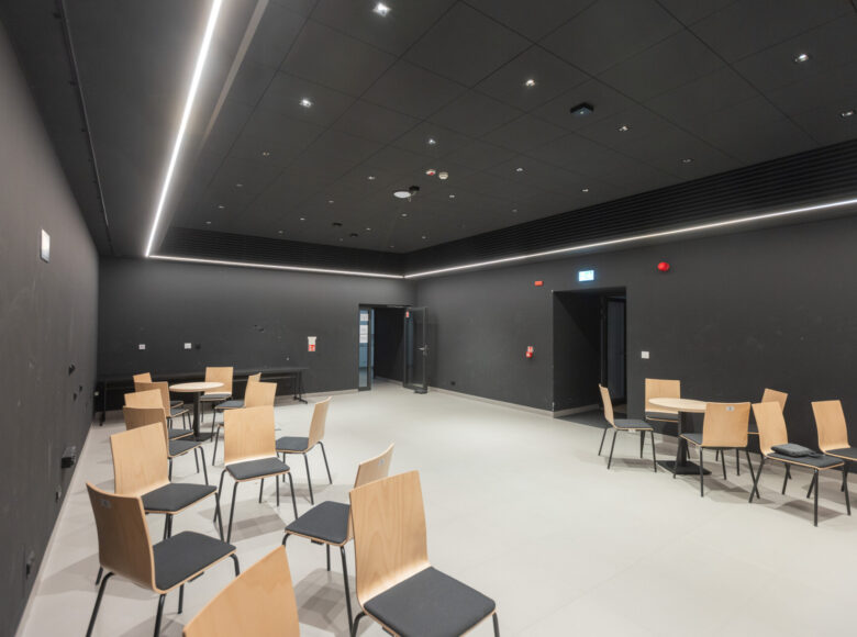 Hol w piwnicach Centrum Kultury w Lublinie. Biała podłoga, czarne ściany i sufit. W holu stoją swobodnie ustawione krzesła i kilka kawiarnianych stolików.