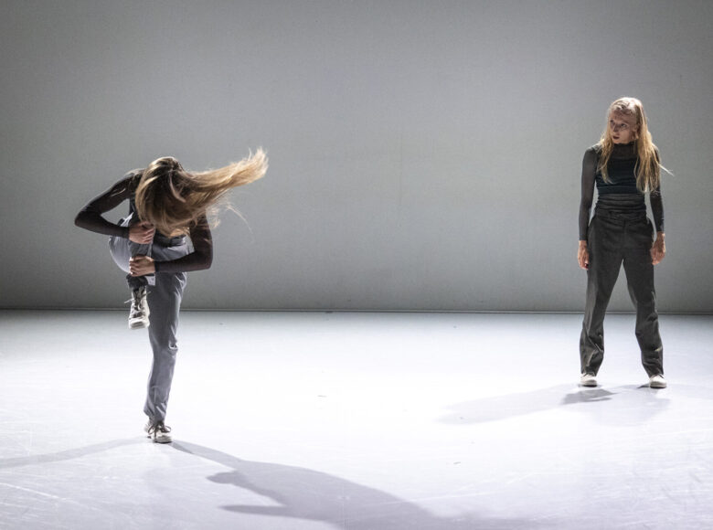 Spektakl teatralny. W jasnym wnętrzu dwie dziewczyny. Pierwsza dziewczyna stoi na jednej nodze, kolano drugiej nogi podciąga do góry. Druga dziewczyna stoi w statycznej pozie i patrzy na pierwszą dziewczynę.