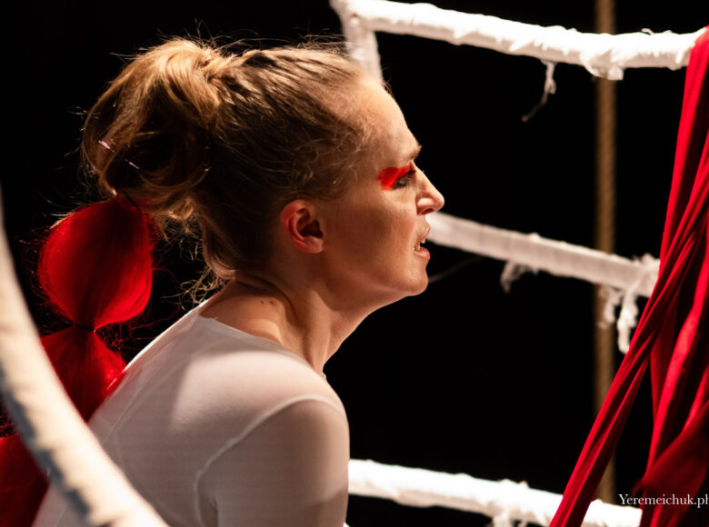 Przedstawienie teatralne. Zbliżenie na twarz kobiety w białej bluzce i z pomalowanymi na czerwono powiekami. Dookoła niej czerwono białe dekoracje. Z tyłu czarne tło.