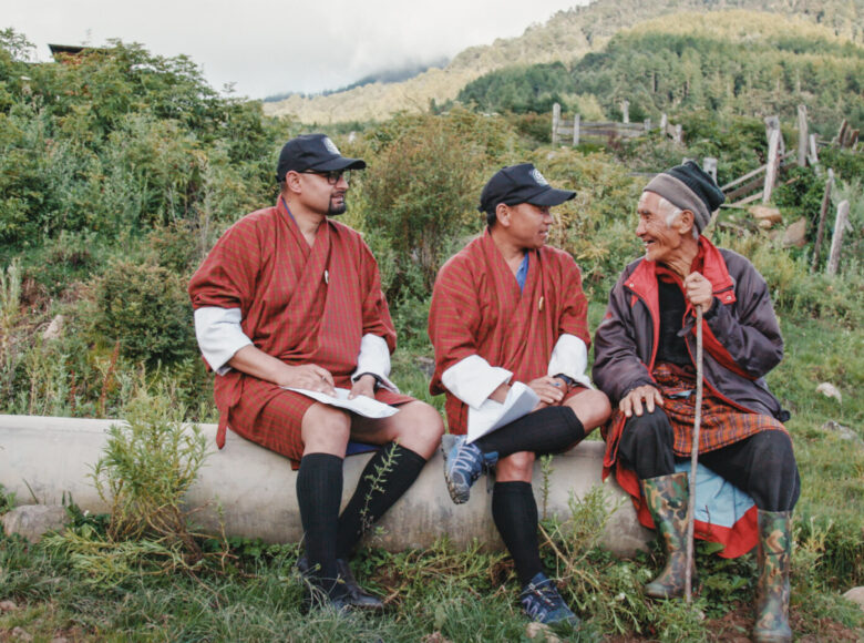Zdjęcie w naturze, dookoła trawa, zarośle, w tle wzgórza. Na pierwszym planie siedzi trzech mężczyzn - młody Europejczyk oraz dwóch Azjatów - młody i stary.