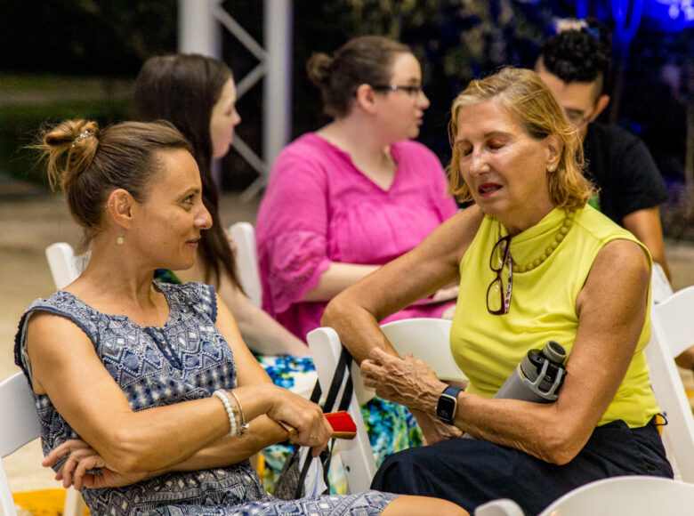 Lato. Na dworze grupa osób siedzących na krzesłach: na pierwszym planie dwie kobiety pogrążone w rozmowie, za nimi trójka młodych ludzi rozmawiających ze sobą.