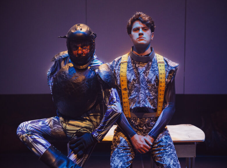 Przedstawienie teatralne. Na scenie na ławce siedzi dwóch mężczyzn w fantazyjnych kostiumach teatralnych w kolorze fioletowym. Jeden z mężczyzn ma na głowie kask hokejowy. Z tyłu jednolite, fioletowe tło.