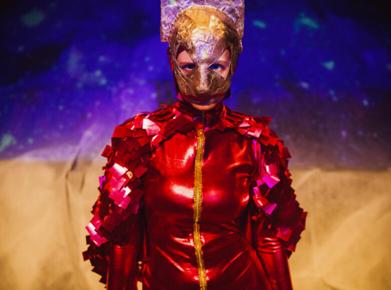 Występ sceniczny. Na scenie zbliżenie na kobietę ubraną w czerwony, błyszczący, obcisły kombinezon i maskę na twarzy. Za nią tło w kolorach żółtym i fioletowym..