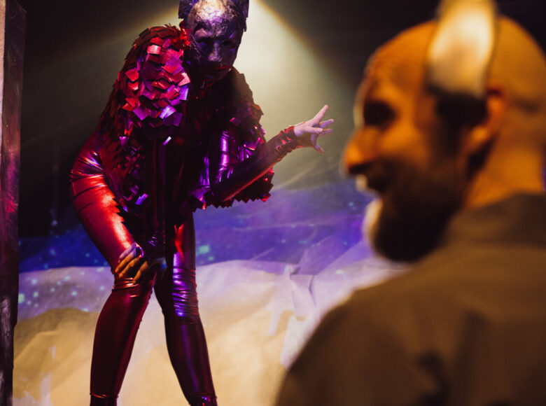 Występ sceniczny. Na scenie kobieta pochylona do przodu, z podniesioną ręką. Ubrana jest w czerwony, błyszczący, obcisły kombinezon i buty na wysokich obcasach. Na twarzy ma maskę.