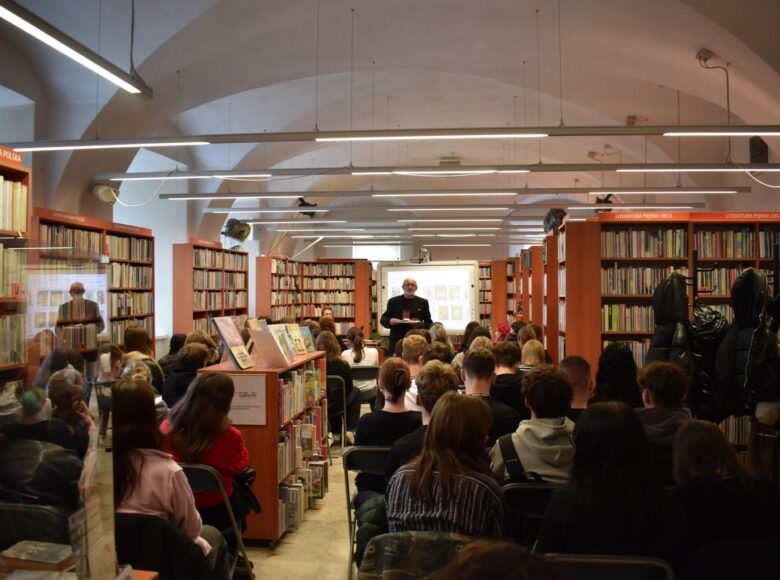 Spotkanie z czytelnikami w Bibliotece Miejskiej w Centrum Kultury w Lublinie. Mężczyzna rozmawia z grupą młodych ludzi siedzących na krzesłach. dookoła regały z książkami.